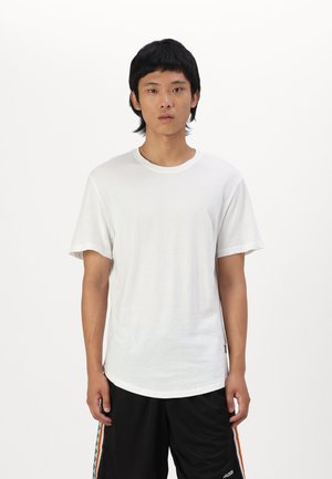 Weißes Baumwoll-T-Shirt mit Rundhalsausschnitt und kurzen Ärmeln, mit lockerer Passform und subtiler Textur. Kombiniert mit schwarzen Shorts.