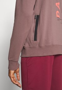 Sweat-shirt bordeaux avec une poche zippée et des poignets côtelés, fabriqué en tissu doux ; présente un texte graphique orange vif sur le devant.