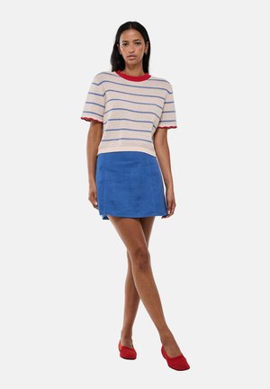 Femme portant un haut rayé à manches courtes avec bordure rouge, une jupe bleue et des chaussures rouges, debout jambes croisées sur un fond uni.