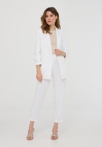 Completo di blazer bianco e pantaloni su misura, abbinato a sandali con tacco a strappo. Tessuto liscio e leggero, linee pulite e dettagli minimalisti.