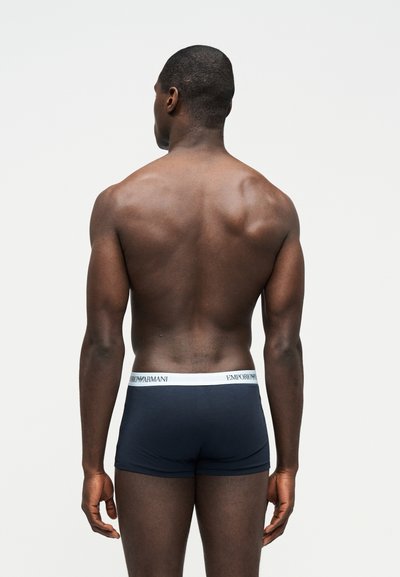 Muskularny czarnoskóry mężczyzna stoi tyłem, ubrany w granatowe bokserki Emporio Armani z białym pasem na talii, na jednolitym tle.