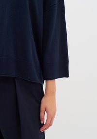 Sötétkék, oversize pulóver bordázott mandzsettákkal és aljjal, puha kötött anyagból. Kéz pihen egy sötét szöveten, a laza szabást bemutatva.
