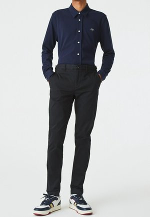 Homme portant une chemise bleu marine à boutons, un pantalon noir et des baskets blanches avec des accents bleu marine et jaune, debout les mains dans les poches.