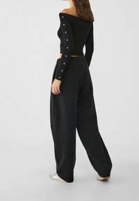 Zwarte off-shoulder ribbed top met lange mouwen en knoopdetails; gecombineerd met losse zwarte broeken en zilveren schoeisel.