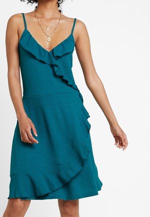 Kobieta w turkusowej sukience bez rękawów z falbanami przy dekolcie w kształcie litery V oraz asymetryczną falbaną na przodzie i na dole spódnicy.