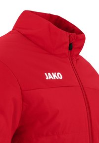 Giacca rossa con colletto alto, realizzata in tessuto testurizzato. Presenta una zip nera e il logo bianco "JAKO" sul lato sinistro.