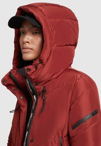 Chaqueta de plumas roja con cuello alto y capucha, que presenta acentos negros, cierre delantero y bolsillos laterales. Textura suave con un diseño acolchado.