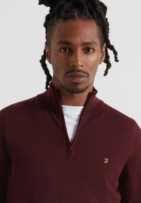 Maglione zip-up borgogna realizzato in tessuto a maglia, con colletto alto e un piccolo logo dorato sul petto. Maglietta bianca visibile sotto.