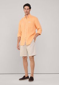 Lichtoranje, langmouw overhemd met knopen en een voorzak, gecombineerd met beige shorts en donkere instapschoenen, staand tegen een neutrale achtergrond.