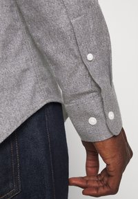 Szara rękaw koszuli o fakturze z detalami guzików w kontrastującym kolorze, zestawiona z ciemnym denimu. Prosty, casualowy design.