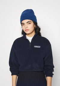 Μπορντό φλις πουλόβερ με φερμουάρ 1/4 και στρογγυλό λογότυπο Adidas. Φορεμένο με ένα μπλε πλεκτό καπέλο. Κοντή σχεδίαση με ελαστικές μανσέτες.