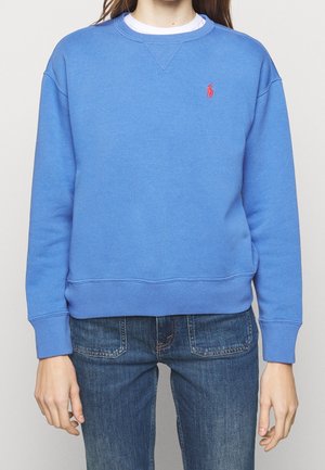 Personne portant un sweat-shirt bleu à manches longues avec un petit logo rouge sur la poitrine gauche et un jean bleu.