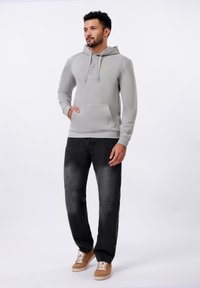 Man wearing a light grey pullover hoodie with buttons, black faded jeans, and brown suede sneakers, standing with one hand in pocket.