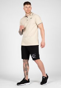 Beige poloshirt met een kraag en bruine accenten, gecombineerd met zwarte shorts met een grafisch ontwerp. Model draagt zwarte sneakers.