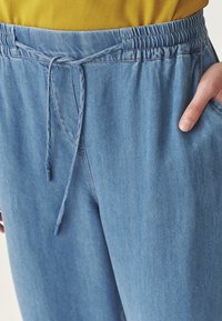 Heledad sinised denim joggers, millel on elastne vöökoht, pael ning küljetaskud. Sile tekstuur koos peente õmblusdetailidega.