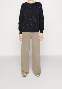 Persona vistiendo un suéter negro de manga larga con puntos blancos, pantalones anchos beige y bailarinas doradas, de pie contra un fondo simple.