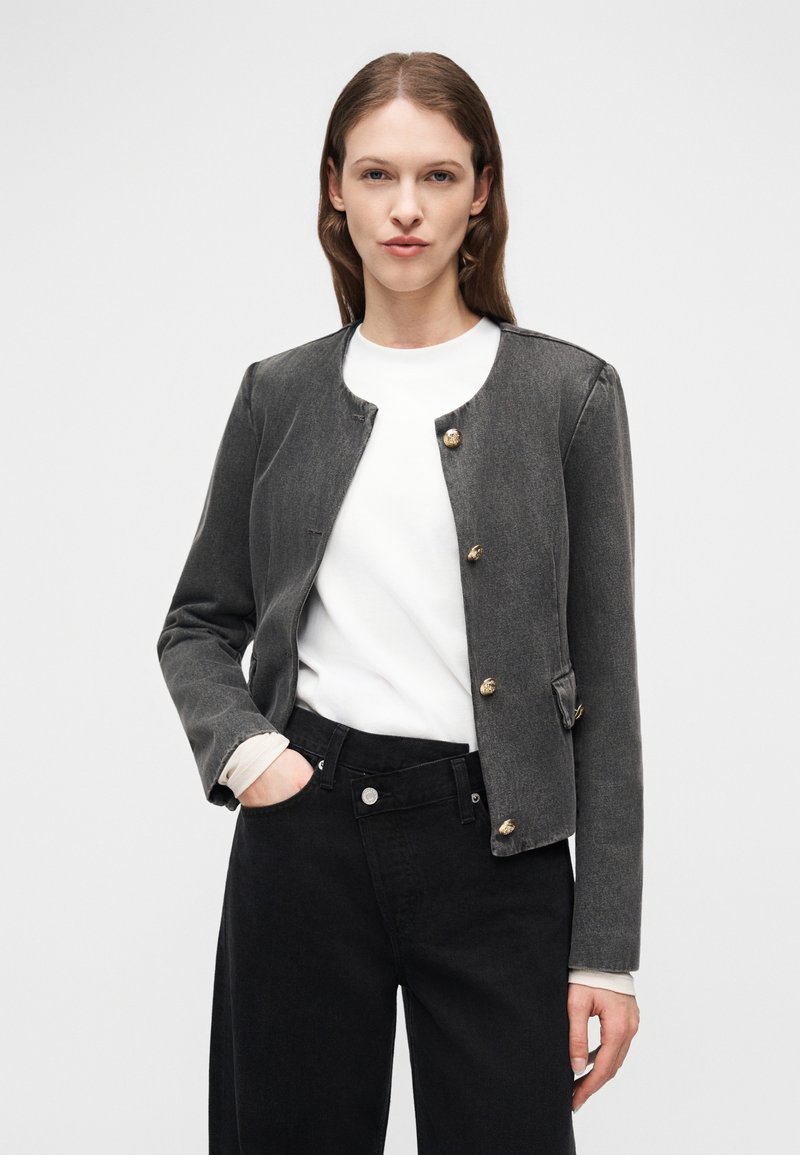Femme portant une veste boutonnée gris foncé par-dessus un haut blanc et un jean taille haute noir, debout avec une main dans la poche.