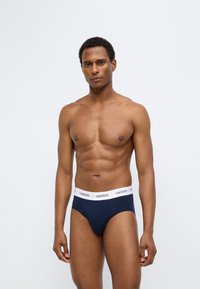 Μπλε σκούρο μαγιό με λευκή ζώνη μέσης που φέρει το λογότυπο της Lacoste. Υφασμάτινη επιφάνεια, εφαρμοστό σχέδιο και μεσαία μέση.