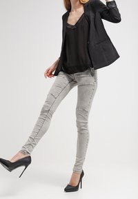 Žena v černém saku, černém topu, šedých skinny džínách a černých vysokých botách, zaujímající dynamickou pózu na jednoduchém pozadí.