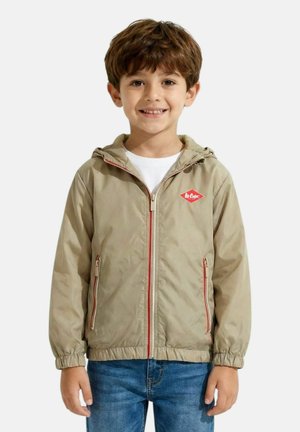 Jeune garçon souriant portant une veste beige zippée avec capuche et logo rouge, chemise blanche, et jean bleu sur un fond uni.