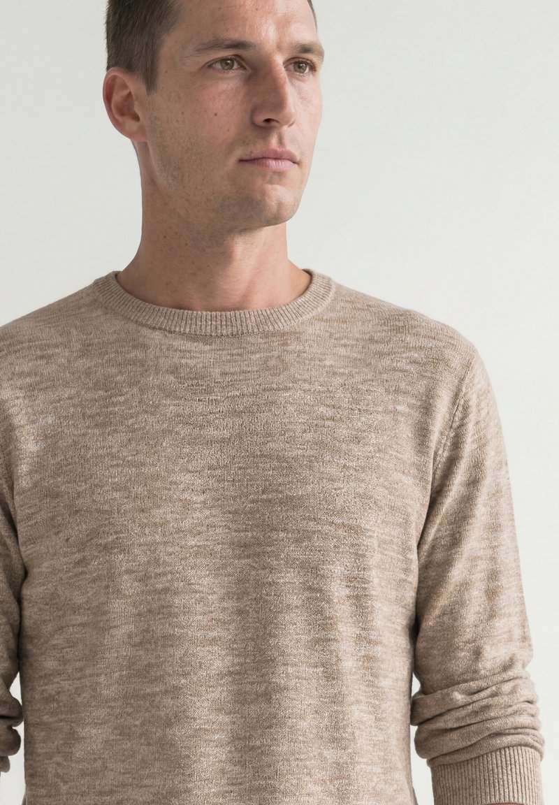 Homme aux cheveux courts et foncés portant un pull beige texturé à manches longues, regardant vers la droite sur un fond uni clair.