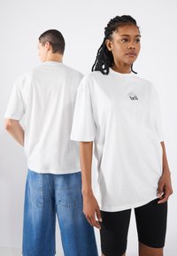 Hvid bomulds t-shirt med korte ærmer og rund halsudskæring, der har et sort grafisk "klokke" design på brystet. Båret med denimshorts.
