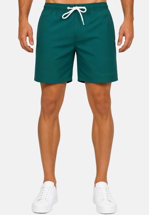 Torso e gambe maschili che indossano pantaloncini da bagno verde scuro con coulisse bianca e sneakers bianche, in piedi davanti a uno sfondo uniforme.