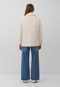 Person mit langen braunen Haaren trägt eine beigefarbene, strukturierte Jacke, weite blaue Jeans und weiße Schuhe und steht vor einem hellgrauen, einfarbigen Hintergrund.