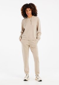 Felpa beige con cappuccio, maniche e orlo a coste, abbinata a pantaloni a sigaretta coordinati. Il modello indossa sneakers di colore chiaro.