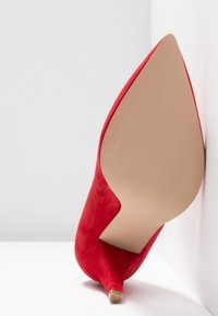 Chaussure à talon haut rouge inclinée, avec la semelle tournée vers l'avant, présentant un bout pointu et une semelle texturée beige sur fond blanc.