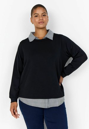 Confident woman with short hair wears black long-sleeve top over striped collared shirt and dark blue jeans, standing against white background.