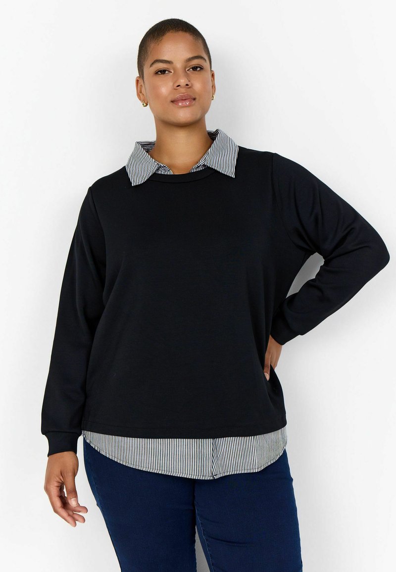 Donna sicura di sé con capelli corti indossa un top nero a maniche lunghe sopra una camicia a righe con colletto e jeans blu scuro, in piedi davanti a uno sfondo bianco.