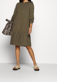 Olijfgroen hoodie jurk met lange mouwen en een gelaagde rok. Gecombineerd met een geruite tas en beige instap-sandalen.