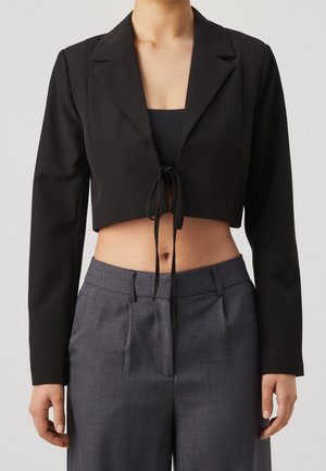 Kvinde iført en sort cropped blazer med knude foran over en sort top, kombineret med højttaljede grå skræddersyede bukser, stående mod en ensfarvet baggrund.