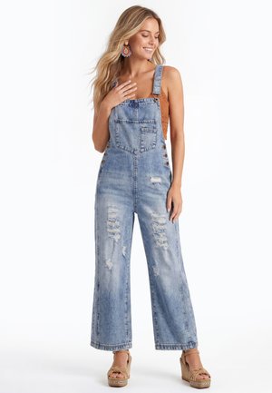 Lachende vrouw met lang blond haar draagt een oranje mouwloze top, versleten blauwe denim tuinbroek, sleehak sandalen en grote kleurrijke oorbellen.