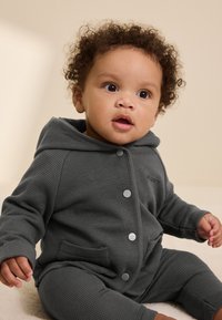 Gray waffle-knit hoodie and pants set for infants, featuring snap buttons, front pockets, and a soft texture. Curly hair visible.