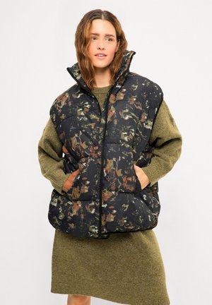 Frau trägt ein locker sitzendes grünes Strickkleid und eine schwarze, gepolsterte Weste mit grün-orange-weißem Camouflage-Muster, Hände in den Westentaschen.