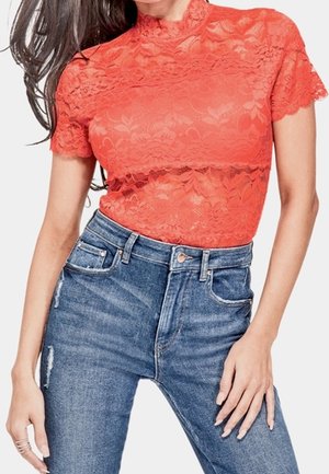 Kvinde iført en tætsiddende, koralfarvet bluse med korte ærmer og blonder, kombineret med højttaljede, let slidte blå jeans.