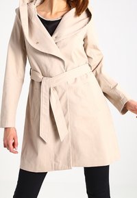 Beige vyötäröllä varustettu trench-coat, jossa on huppu, minimimäiset napit ja sivutaskut. Takissa on sileä kangas ja istuva malli.