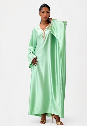 Femme portant une longue robe en satin vert menthe avec une bordure brodée beige, des boucles d'oreilles créoles dorées et des sandales à talons hauts dorées, posant la main sur la tête.