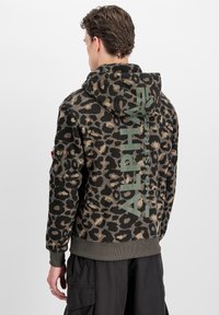 Giovane con felpa leopardata con grande scritta verticale "Alpha Industries" sulla schiena, abbinata a pantaloni cargo neri.