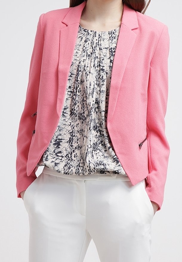 Femme portant un blazer rose par-dessus un chemisier à motifs et un pantalon blanc, mains dans les poches, sur un fond uni.