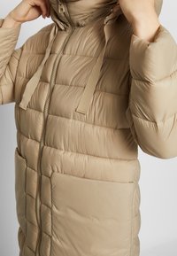 Béžová péřová bunda s prošívaným designem, zipovým zapínáním vpředu a kapucí. Má hladkou texturu a dvě přední kapsy.