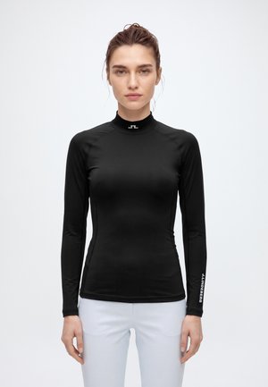 Una mujer posa con un top negro ajustado de manga larga y cuello alto, y pantalones blancos, sobre un fondo liso y claro.
