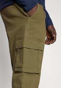 Pantalones cargo de color verde oliva, hechos de una tela resistente, con múltiples bolsillos y detalles cosidos. Tienen un corte ajustado y una cintura cómoda.