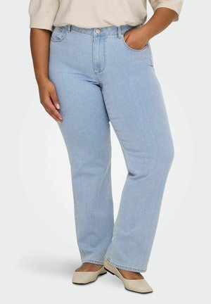 Asmuo dėvi šviesiai mėlynas tiesias džinsines kelnes, smėlio spalvos lakuotus batelius ir kreminę megztinę, stovi viena ranka kišenėje, kita atsipalaidavusi.