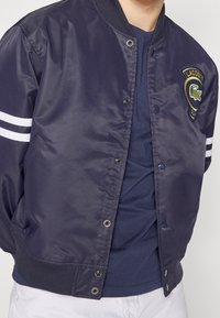 Námořnická modrá bomber bunda s lesklým povrchem, bílé pruhy na rukávech, vyšívané logo, patentky a pružné manžety a lem.