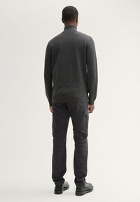 Donkergrijze coltrui met een korte hals, gecombineerd met donkere cargobroek, die een gestructureerde stof en discrete zakken toont, gedragen met zwarte schoenen.