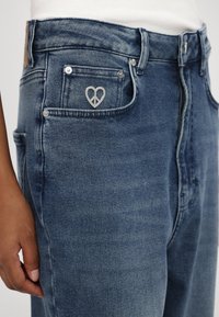 Spijkerbroek in medium blauw met lichte slijtage. Heeft een vredesteken hartembleem op de voork pocket en zilveren hardware accenten.