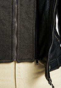 Musta nahkatakki, jossa on harmaa huppari sisällä, varustettuna kahdella vetoketjulla, teksturoidulla kankaalla ja beigeillä housuilla alla.